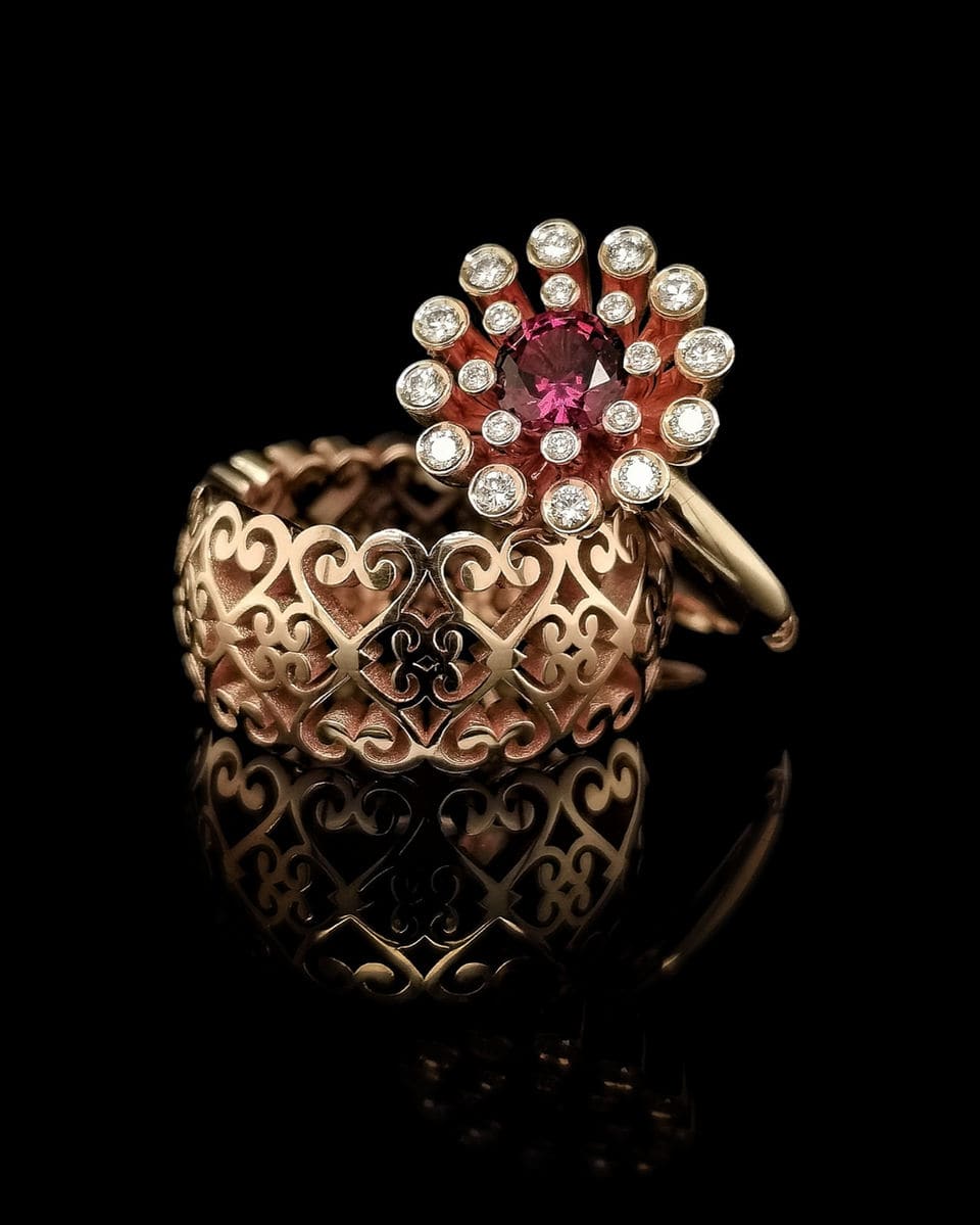 Ring met een roze steen in een krans van briljanten, naast een band met hartvormig krulwerk.